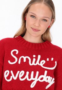 Roter Strickpullover mit weißem Text "Lächle jeden Tag", runder Ausschnitt und strukturiertem Stoff. Runde rote Ohrringe ergänzen den Look.