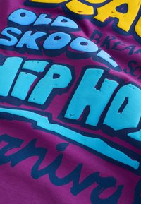 Élénk lila szövet, nagy, világoskék és sárga vastag betűkkel, amelyeken részben az "Old Skool Hip Hop" felirat olvasható, valamint kisebb fekete szöveg a háttérben.