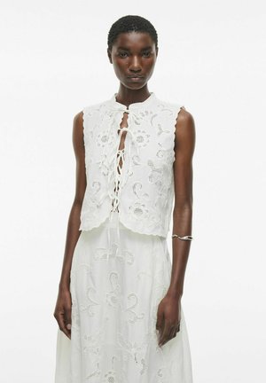 Haut sans manches blanc avec des accents en dentelle, ourlet festonné et détail de nœud à l'avant ; présente une broderie florale texturée sur l'ensemble.