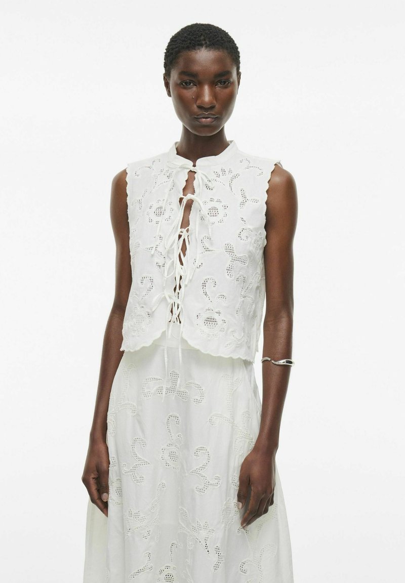Haut sans manches blanc avec des accents en dentelle, ourlet festonné et détail de nœud à l'avant ; présente une broderie florale texturée sur l'ensemble.