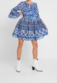 Modré šaty s vzorem, které mají výstřih ve tvaru klíčové dírky, vrstvenou sukni a rozšířené rukávy; vyrobené z lehkého materiálu s detailním tiskem.