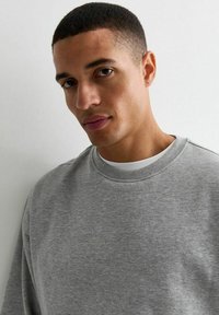 Sudadera gris de cuello redondo en una tela suave, con un acabado texturizado y cuello acanalado, combinada con una camiseta blanca debajo.