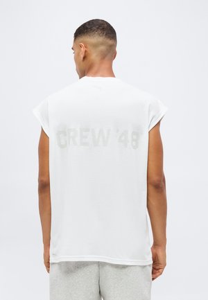 Mann mit kurzen Haaren trägt ein weißes ärmelloses Shirt mit dem Text "CREW '48" auf dem Rücken und hellgraue Shorts, steht mit dem Rücken zur Kamera.