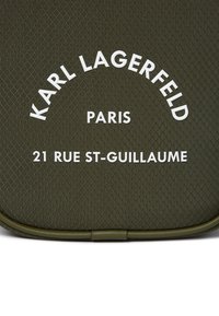 Superficie di colore verde oliva, testurizzata, con testo bianco in rilievo: "KARL LAGERFELD PARIS 21 RUE ST-GUILLAUME" in un design ad arco.
