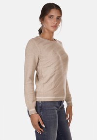 Jersey de punto beige con patrón texturizado diagonal, cuello redondo, mangas largas y dobladillo acanalado. Llevado con jeans oscuros.
