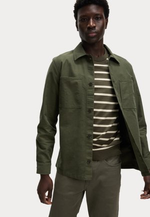 Olive green jacket with two chest pockets, button closure, worn over a striped sweater. Matched with slim-fit green pants.