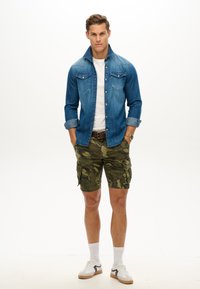 Jeans skjorte med knapper, hvid t-shirt, camouflage cargo shorts, hvide sneakers med sorte detaljer og hvide sokker. Model poserer med hænderne i lommerne.