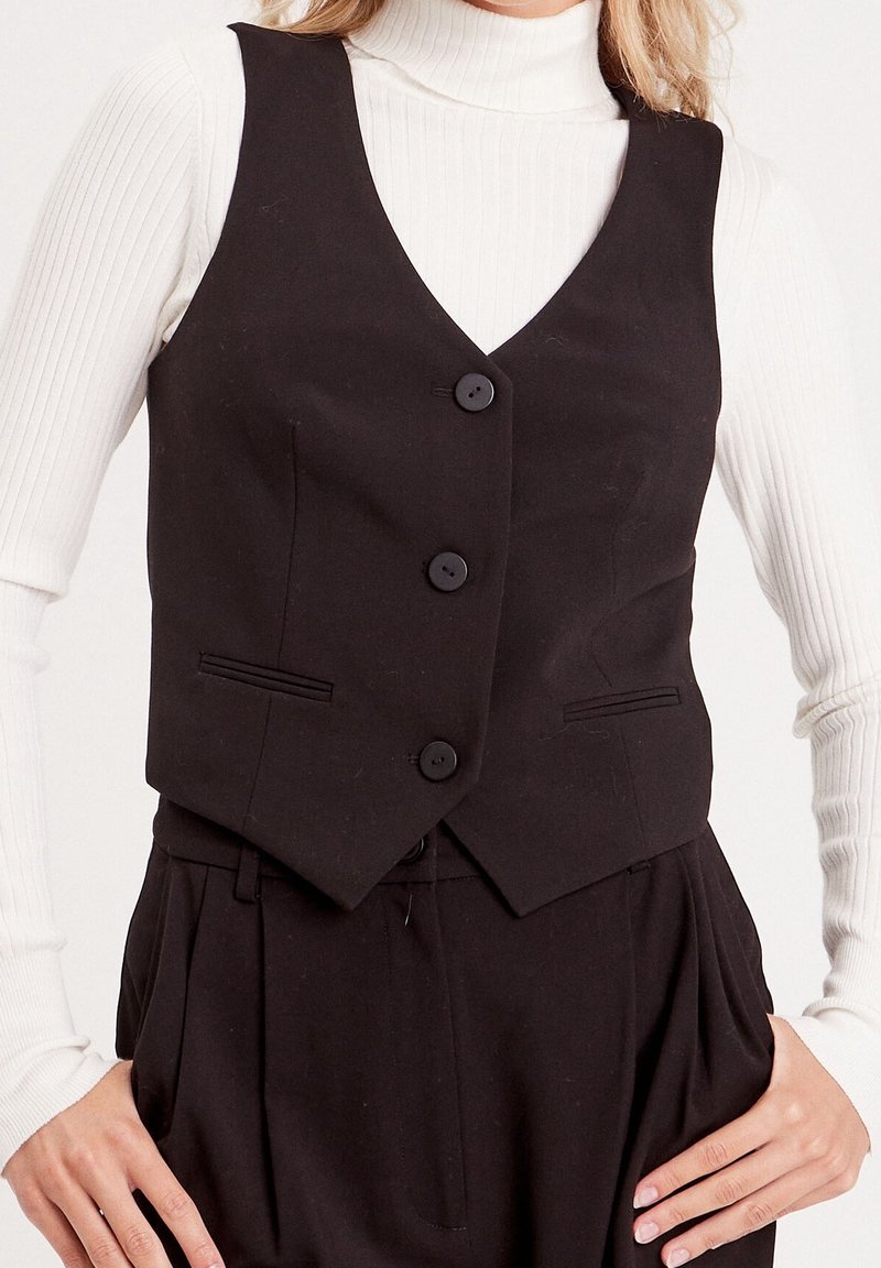 Zwarte bodywarmer met een getailleerd ontwerp, voorzien van drie knopen en voorkantenzakken. Gemaakt van gladde stof en gecombineerd met een witte geribbelde coltrui.