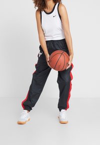 Czarne sportowe spodnie z czerwonymi paskami po bokach i logo, w połączeniu z białym topem na ramiączkach i białymi trampkami. Osoba trzyma piłkę do koszykówki.