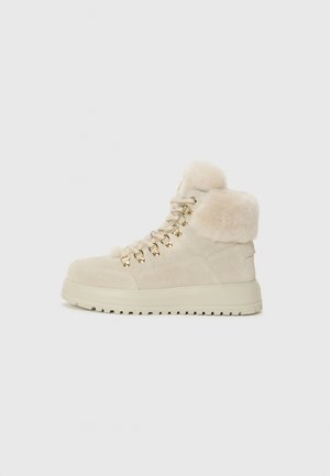 Bottines montantes beiges avec col en fausse fourrure, œillets en métal doré et semelle épaisse, sur fond blanc uni.