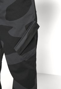Pantalon cargo camouflage gris foncé et noir avec une poche à rabat velcro sur la cuisse latérale, vu sur fond blanc.