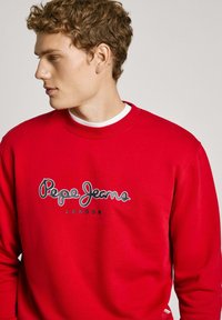 Rdeča jope iz bombaža, z okroglim izrezom in rebrastimi manšetami. Na sredini prikazuje logotip "Pepe Jeans" v beli in črni barvi. Gladka tekstura.