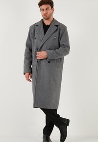 Uomo vestito con un lungo cappotto di lana grigio doppiopetto, pantaloni neri e stivali neri, in piedi con una gamba incrociata davanti.