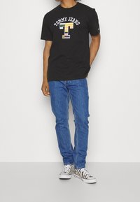 Svart bomull t-shirt med "TOMMY JEANS" logotyp i flerfärg ovanför ett stiliserat bokstav "T", bärs med blå denimjeans och mönstrade sneakers.