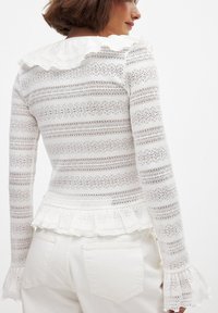 Haut à manches longues blanc avec un col ondulé et des poignets évasés. Présente un motif rayé texturé et un ourlet en peplum.