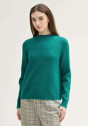 Türkisfarbener gerippter Pullover mit hohem Kragen und langen Ärmeln, aus strukturiertem Material, kombiniert mit karierten Hosen.