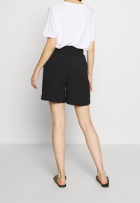 Shorts noirs avec une ceinture élastique, présentant une coupe décontractée et un tissu lisse. Associés à une chemise blanche ample et des sandales.