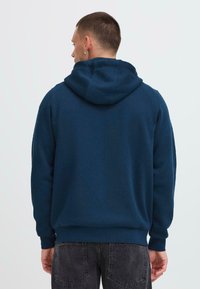 Tengerészkék fleece kapucnis pulóver texturált felülettel, nagy kapucnival és bordázott mandzsettákkal. Szürke, elhasználódott farmerekkel viselve, amely laza szabású.