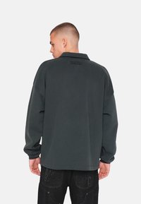 Felpa oversize grigio scuro con colletto piegato e maniche lunghe; presenta un logo tonale sulla parte alta della schiena; texture liscia.