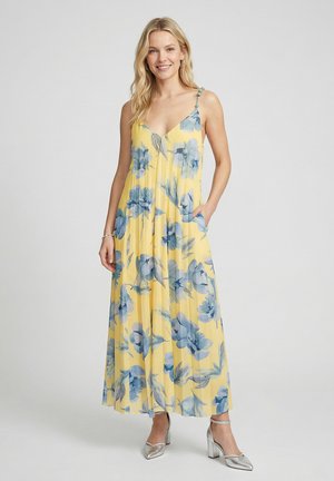 Vrouw draagt een mouwloze gele maxi-jurk met blauw bloemmotief en zilveren hakken, staat met één hand in de zak en lacht.