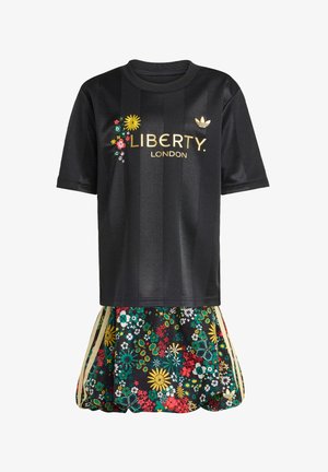 Μαύρη αθλητική φανέλα με χρυσή επιγραφή "LIBERTY LONDON" και διακοσμητικά με λουλούδια, σε συνδυασμό με φούστα με λουλουδένιο μοτίβο που διαθέτει πράσινες, κόκκινες και κίτρινες λεπτομέρειες.