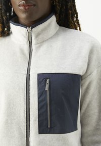 Hellgraue Fleecejacke mit einer marineblauen Reißverschlusstasche auf der Brust und einem Kragen. Verfügt über einen zentralen Reißverschluss und strukturierten Stoff.