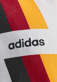 Κοντινή λήψη υφάσματος με μαύρο λογότυπο «adidas» και έντονες κάθετες ρίγες σε μαύρο, κόκκινο και κίτρινο σε λευκό φόντο.