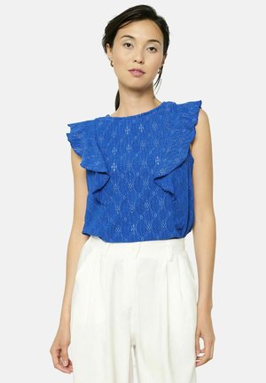 Haut sans manches bleu avec des accents de volants, tissu texturé présentant des motifs en diamant, associé à un pantalon blanc taille haute.