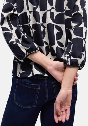 Personne portant un haut à manches longues à motifs noir et blanc et un jean bleu foncé, les mains croisées dans le dos, montrant une bague en argent.