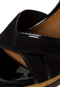 Sandalia negra que presenta una combinación de charol brillante y suave ante. Diseño con recortes y forro interior marrón, además de una suela texturizada.