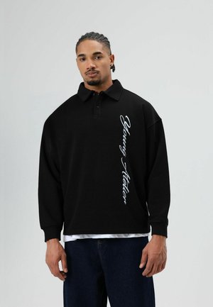 Zwart poloshirt met een ontspannen pasvorm, lange mouwen en een witte cursieve "Young Atelier"-print langs de zijkant. Gedragen over een wit T-shirt.