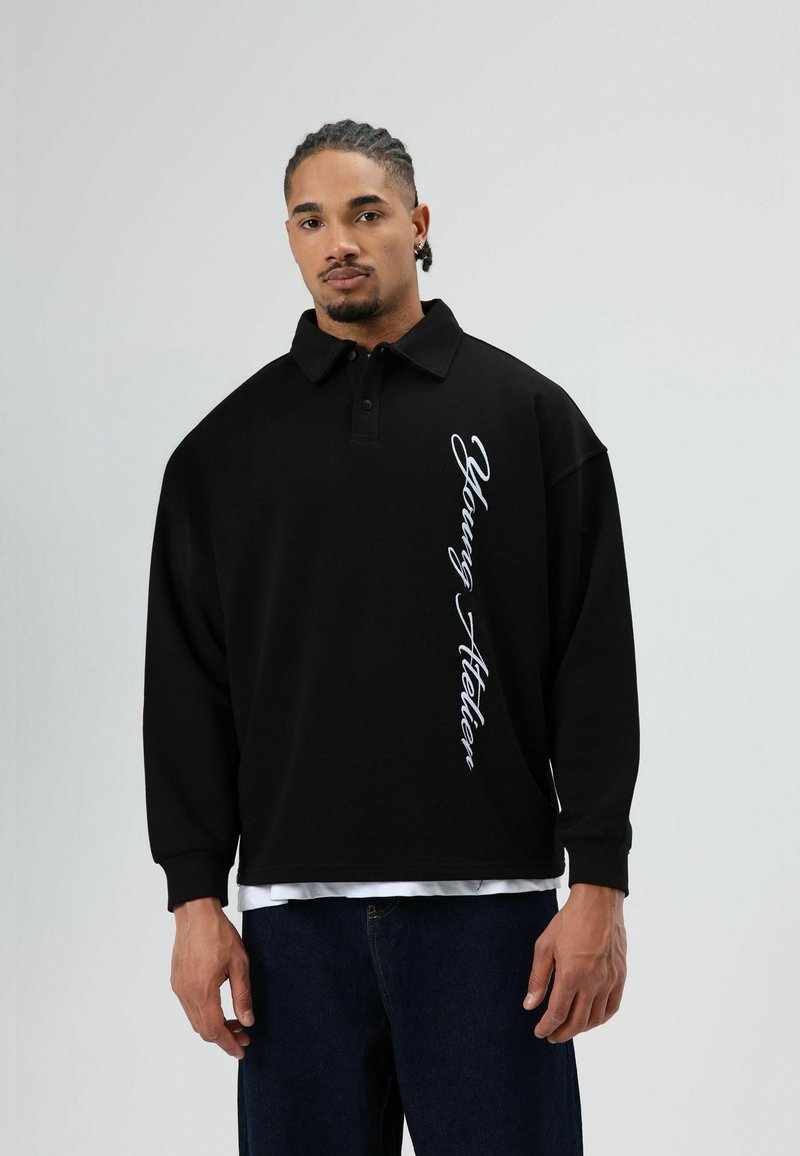 Zwart poloshirt met een ontspannen pasvorm, lange mouwen en een witte cursieve "Young Atelier"-print langs de zijkant. Gedragen over een wit T-shirt.