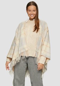 Poncho à carreaux en crème clair, gris et pêche avec un ourlet à franges ; porté sur un pull en beige texturé et un jean gris clair.