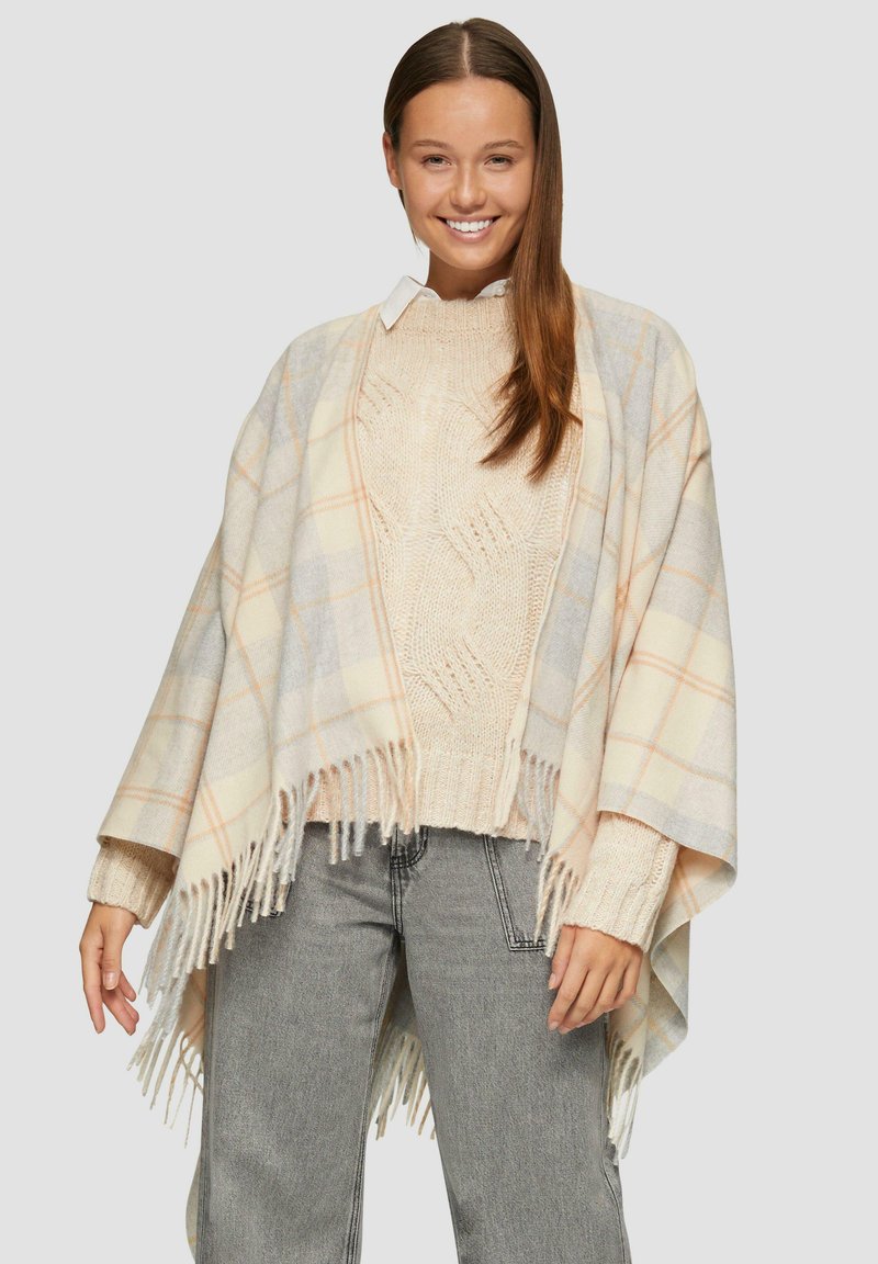 Poncho à carreaux en crème clair, gris et pêche avec un ourlet à franges ; porté sur un pull en beige texturé et un jean gris clair.