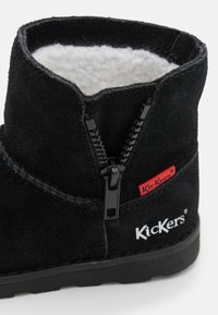 Botte en suède noire avec doublure en fourrure blanche, fermeture éclair sur le devant et logo Kickers rouge. Semelle en caoutchouc noir plat. Hauteur de cheville courte.