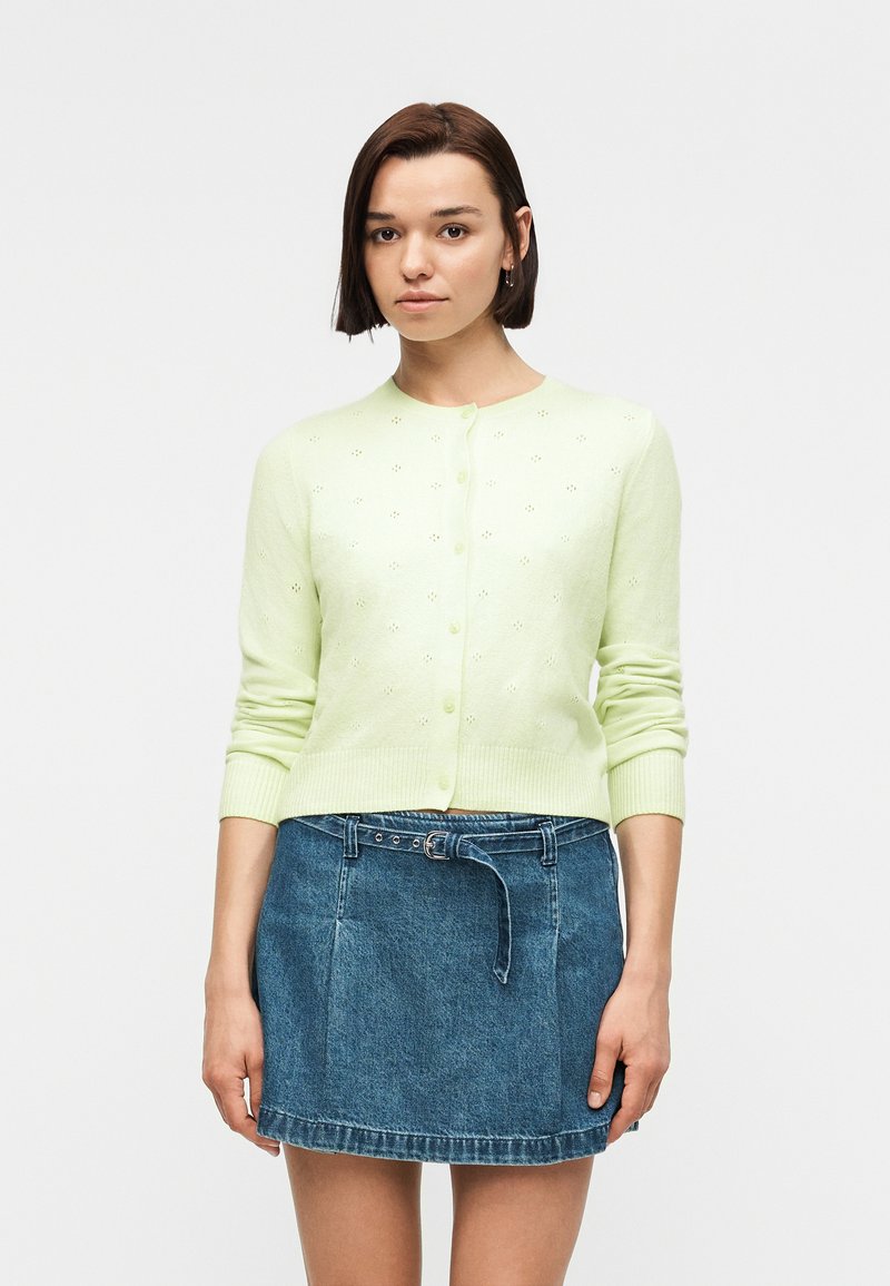 Femme aux cheveux courts bruns portant un cardigan vert clair boutonné et une jupe en jean bleue ceinturée, debout devant un fond blanc.