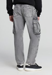 Pantaloni cargo in denim grigio con finitura leggermente sbiadita, dotati di tasche laterali, polsini elastici e vestibilità rilassata. Sneakers nere completano l'outfit.