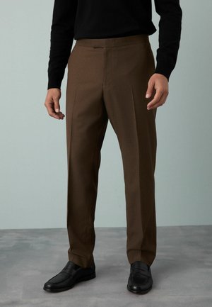Pantalon marron ajusté avec une texture lisse et un devant plissé, associé à des mocassins noirs, sur un fond atténué.
