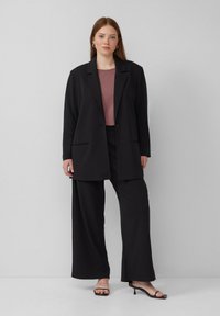 Schwarzer maßgeschneiderter Blazer mit zwei Fronttaschen, kombiniert mit einer lockeren schwarzen Hose. Darunter ein figurbetontes mauvefarbenes Oberteil. Die Accessoires umfassen Creolen.