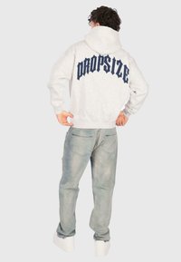 Osoba v svetle šedé mikině s nápisem "DROPSIZE" na zádech, vybledlých modrých džínách a bílých teniskách, stojící s rukama u pasu.