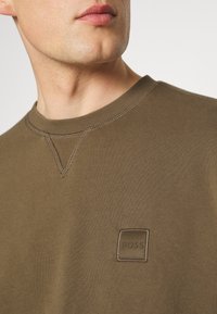 Manlig modell som bär en brun tröja med rund hals och en liten broderad "BOSS"-logotyp på bröstet, visad från halsen till övre torso.