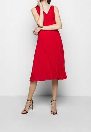 Robe midi plissée rouge avec décolleté en V, sans manches et silhouette fluide. Associée à des sandales noires à talons hauts et fines brides.