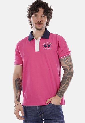 Uomo con capelli ricci che indossa una polo rosa con colletto navy e loghi, che mostra tatuaggi dettagliati su entrambi gli avambracci, posando con la mano in tasca.