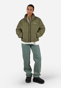 Olivově zelená péřová bunda s vysokým límcem, blokovým prošíváním, předním zipem, nošena přes bílý top, v kombinaci s volnými světle modrými džíny a černými botami.