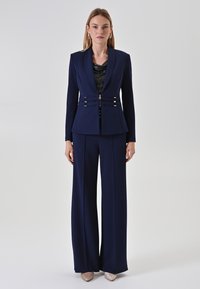 Marineblauer maßgeschneiderter Anzug mit einem taillierten Blazer mit Metallakzenten und weit geschnittenen Hosen. Texturiertes schwarzes Oberteil darunter. Neutrale Absätze.