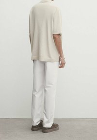 Beige korte mouwen gebreide polo met geribbelde zoom, gecombineerd met witte wijde broek en bruine instapschoenen, gezien van de achterkant.