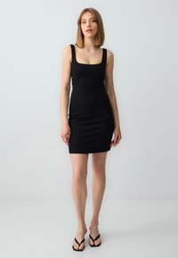 Schwarzes, figurbetontes Kleid mit quadratischem Ausschnitt, breiten Trägern, glatter Textur und knielangem Design. Getragen mit schwarzen Flip-Flops.