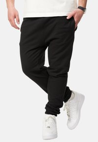 Black jogger pants with a relaxed fit, elastic cuffs, and side pockets, paired with white sneakers. Smooth cotton blend fabric.