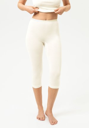 Vrouw draagt witte aansluitende korte legging en een witte top, tilt de top iets op om de buik te laten zien, staat op blote voeten tegen een effen achtergrond.