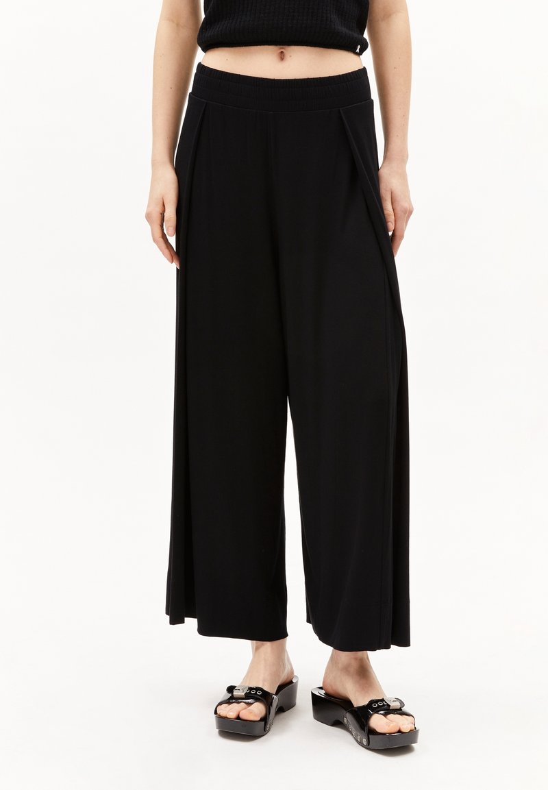 Pantalons larges noirs en tissu lisse avec une taille élastique, présentant des plis pour plus de texture, associés à des sandales à plateforme noires brillantes.